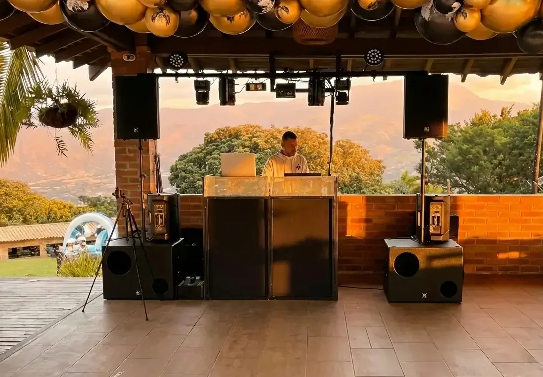 Montaje profesional de sonido, iluminación y cabina de DJ (Andres DJ) en la terraza cubierta de una finca con vista a las montañas de Medellín durante el atardecer, preparado para un evento empresarial privado.